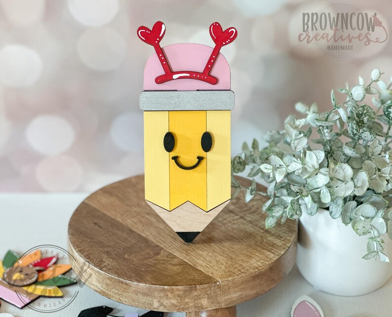 May include: A wooden pencil-shaped decoration with a smiling face, yellow body, and black tip. It has a pink top with red heart-shaped accents and the text "browncow creatives handmade". The pencil sits on a wooden stand.