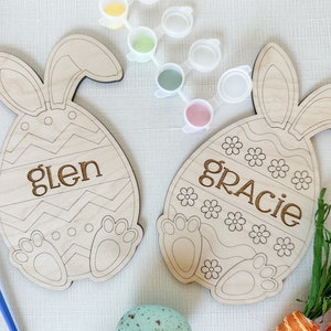 Op de afbeelding: Twee onafgewerkte houten paaseieren in de vorm van een konijn met de namen "Glen" en "Gracie" erop geschilderd. De eieren zijn omringd door verfpotjes en kwasten.