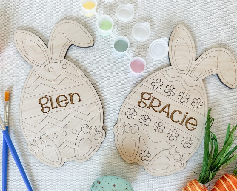 Op de afbeelding: Twee onafgewerkte houten paaseieren in de vorm van een konijn met de namen "Glen" en "Gracie" erop geschilderd. De eieren zijn omringd door verfpotjes en kwasten.
