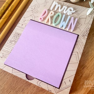 May include: A wooden desk organizer with a pastel pink sticky note and the text "Mrs. Brown" in colorful letters. The organizer features a pencil, ruler, calculator, and other school supplies.