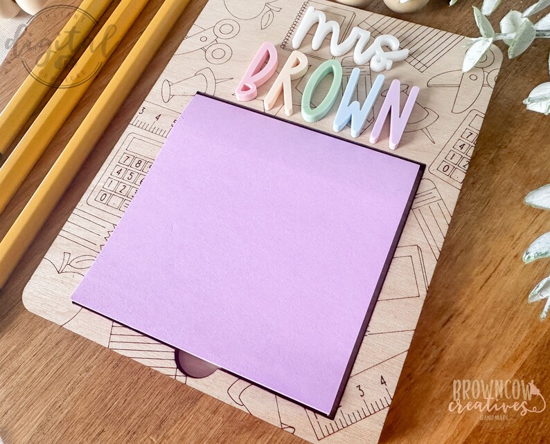 May include: A wooden desk organizer with a pastel pink sticky note and the text "Mrs. Brown" in colorful letters. The organizer features a pencil, ruler, calculator, and other school supplies.
