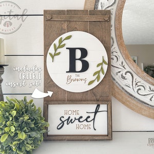 May include: A wooden sign with a white background and a green wreath with the letter "B" in the center. The text "The Browns" is below the letter. The sign is attached to a larger wooden frame with the text "Home Sweet Home" on a smaller sign below it.