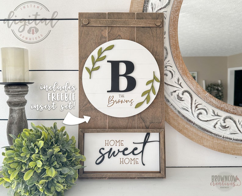 May include: A wooden sign with a white background and a green wreath with the letter "B" in the center. The text "The Browns" is below the letter. The sign is attached to a larger wooden frame with the text "Home Sweet Home" on a smaller sign below it.