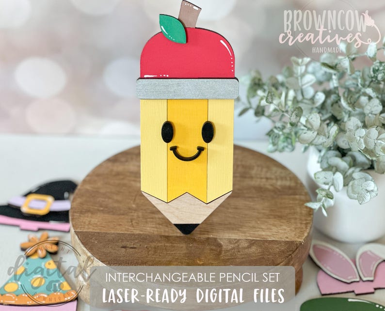 May include: A wooden pencil-shaped decoration with a smiling face, topped with a red apple and green leaf. The pencil is yellow with a gray band and black tip. Text on the image reads "INTERCHANGEABLE PENCIL SET" and "LASER-READY DIGITAL FILES."