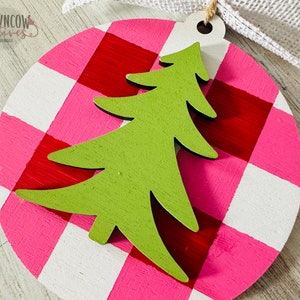 May include: A round, pink and white gingham ornament with a green wooden Christmas tree cutout. The ornament has a hole at the top for hanging.