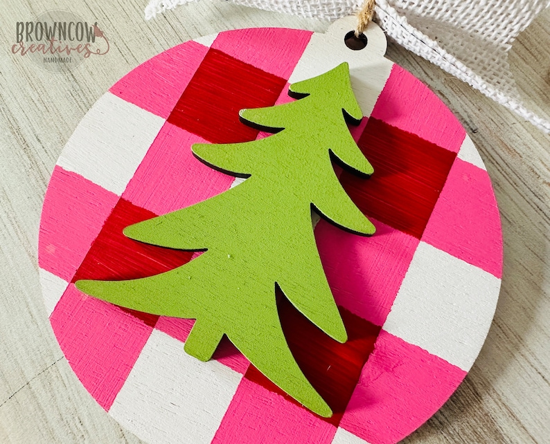 May include: A round, pink and white gingham ornament with a green wooden Christmas tree cutout. The ornament has a hole at the top for hanging.