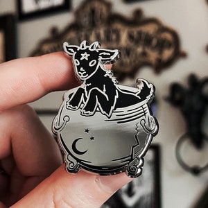 May include: A silver-colored pin featuring a black goat inside a cauldron. The cauldron has a crescent moon and star design. The goat has a star on its forehead. The pin is held by a person.