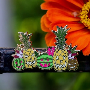 May include: Five enamel pins featuring pineapple and watermelon jack-o'-lantern faces. The pins are arranged in a row on a black surface in front of a bouquet of orange, purple, and yellow flowers.