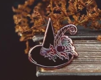 May include: A black witch hat enamel pin with a decorative design, including a red bow and leaves. The pin is set against a backdrop of dried foliage and an antique book, creating a Halloween or autumn theme.