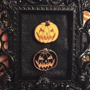 Peut inclure: Un cadre photo noir et orné contient deux épingles en émail sur le thème d'Halloween. L'épingle supérieure est une citrouille jaune avec un visage sculpté, et l'épingle inférieure est une citrouille noire avec un contour cuivré. Le cadre est tenu par une main.