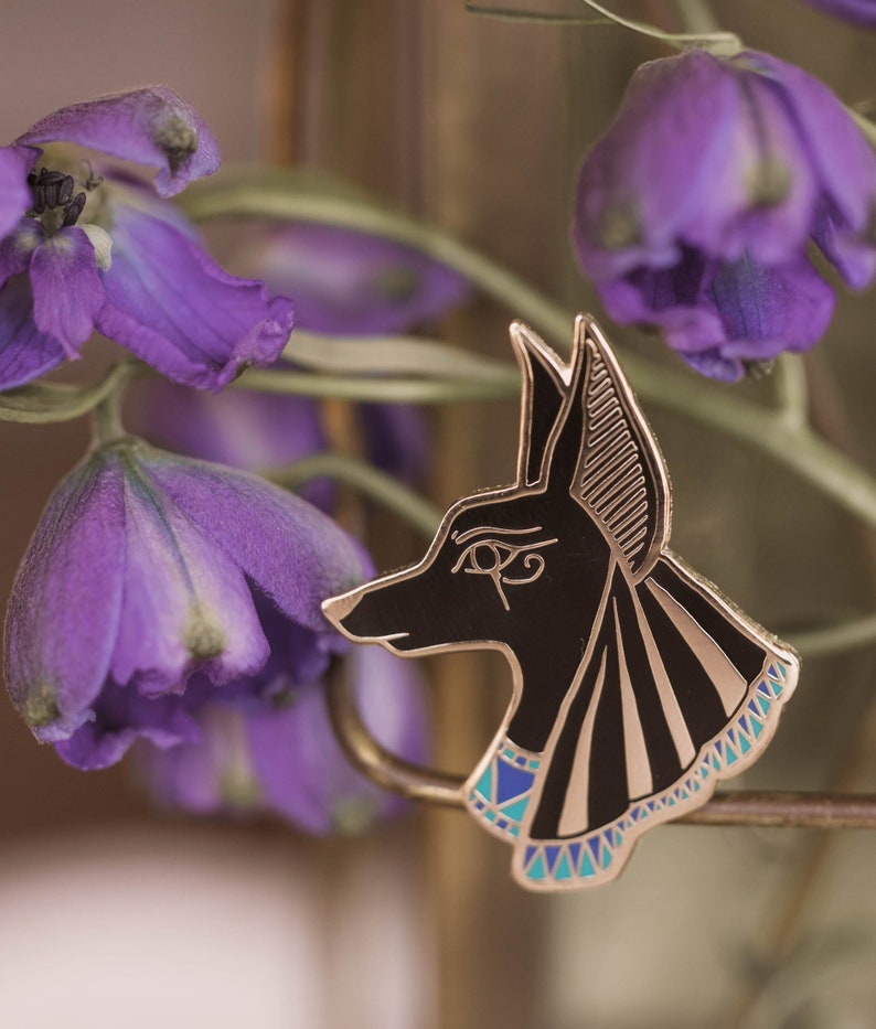 May include: A black and gold enamel pin featuring the profile of Anubis, the Egyptian god of the afterlife. The pin has a detailed design with a gold outline, black body, and blue and gold accents. The pin is set against a blurred background of purple flowers.