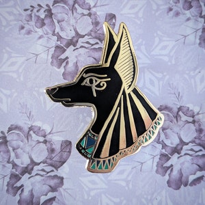 May include: A gold-colored Anubis pin with black enamel details. The pin features the head of the Egyptian god Anubis, with an eye of Horus symbol. The pin has a gold border and teal and gold accents on the collar.