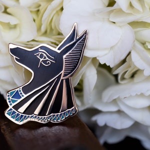 May include: A black and gold Anubis pin with blue and white accents. The pin features the head of the Egyptian god Anubis, with intricate details and a polished finish. The pin is set against a backdrop of white flowers.