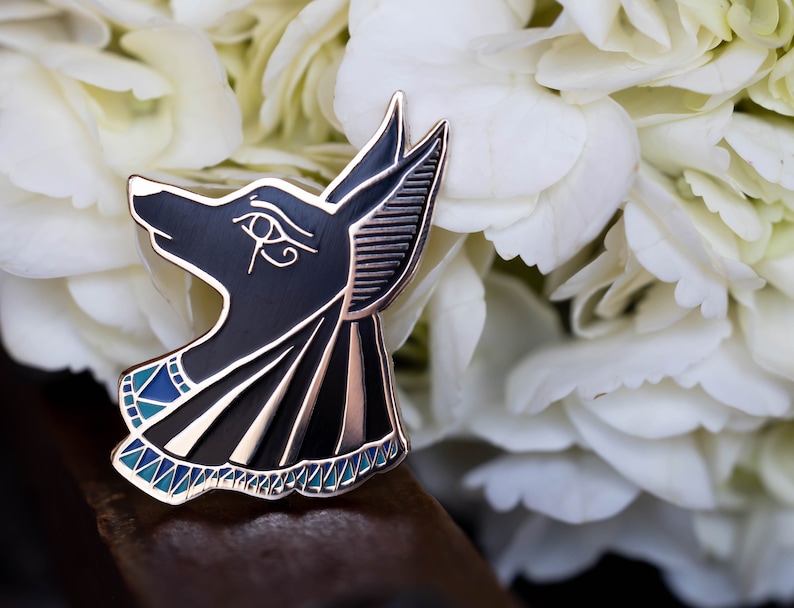 May include: A black and gold Anubis pin with blue and white accents. The pin features the head of the Egyptian god Anubis, with intricate details and a polished finish. The pin is set against a backdrop of white flowers.