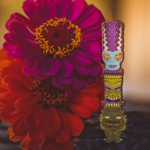 Może przedstawiać: Kolorowa szpilka emaliowana przedstawiająca totem tiki z trzema r&oacute;żnymi twarzami. Totem jest zielony, niebieski, fioletowy i ż&oacute;łty. Szpilka znajduje się przed r&oacute;żowym i pomarańczowym kwiatem.