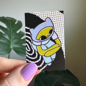 May include: A small, rectangular sticker featuring a cartoon character with a yellow face, blue hood, and blue and yellow arms. The background is black with white dots and swirls. The sticker is held by a hand with purple nail polish.