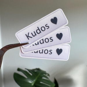 May include: Three rectangular stickers with the word "Kudos" and a black heart symbol. The stickers are white with black text and a black heart. The stickers are being held by a pair of tweezers.