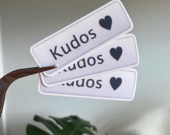 May include: Three rectangular stickers with the word "Kudos" and a black heart symbol. The stickers are white with black text and a black heart. The stickers are being held by a pair of tweezers.
