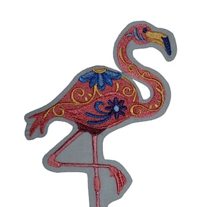 May include: Pink flamingo embroidered patch with gold and blue floral details. The flamingo is standing on one leg with its head turned to the side.