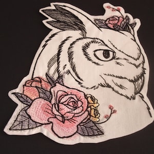May include: Embroidered patch featuring a white owl with black detailing, surrounded by pink and yellow roses. The owl has a serious expression and its eyes are black.