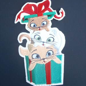 May include: Three embroidered kittens peeking out of a green and red striped gift box with a red bow on top.