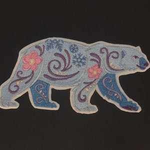 Op de afbeelding: Een blauwe geborduurde ijsbeer met bloemen- en sneeuwvlokmotieven. De beer loopt en heeft een witte omtrek.