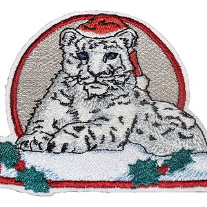May include: Embroidered iron-on patch featuring a white snow leopard wearing a red Santa hat. The leopard is sitting on a white snowy surface with green holly leaves and red berries. The patch is surrounded by a red border.