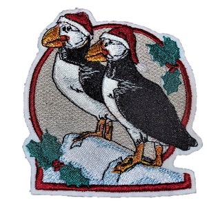 Può includere: Due pulcinelle di mare ricamate con cappelli di Babbo Natale, appollaiate su una roccia innevata con foglie di agrifoglio. Le pulcinelle di mare sono in una cornice circolare rossa e bianca.