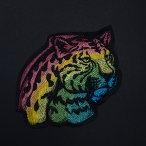May include: Rainbow colored embroidered cheetah head patch. The patch is made of fabric and has a black outline.