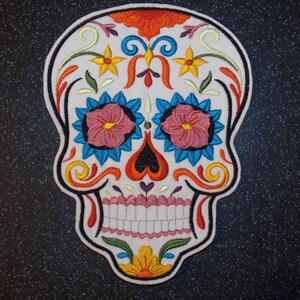 May include: A white embroidered sugar skull with colourful floral details. The skull has a pink and blue flower design for the eyes, a red heart shape for the nose, and a white smile with teeth.