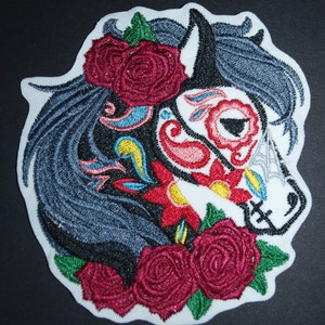 May include: Embroidered iron-on patch featuring a black and white horse with a sugar skull design. The horse is adorned with red, pink, yellow, and blue flowers. The patch is surrounded by green leaves and red roses.