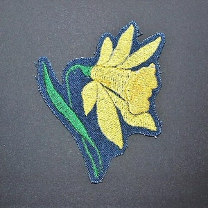 Puede incluir: Una flor de narciso bordada de color amarillo con hojas verdes sobre un fondo de tela azul oscuro.