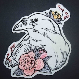 May include: White embroidered crow with black outlines, perched on a bed of pink and orange roses with green leaves. The crow is facing left and has a detailed beak and eye.