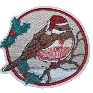 May include: Iron-on patch featuring an embroidered robin wearing a Santa hat and perched on a branch with holly leaves. The robin is brown and white with a red breast and a white belly. The patch is circular with a red and white border.