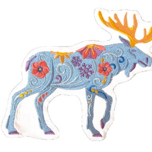 以下が含まれることがあります： 白い背景に立っている、花と雪の結晶のデザインが施された青い刺繍のヘラジカ。