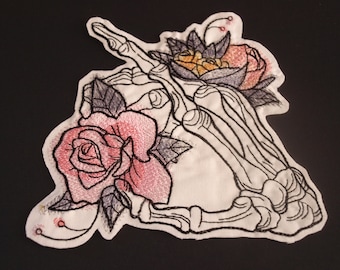 May include: Embroidered patch featuring a skeletal hand holding two roses. One rose is pink and the other is yellow. The patch is on a white background.