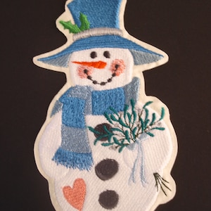 Könnte beinhalten: Weisse Filz-Schneemann-Applikation mit einem blauen Hut, einem blauen Schal und grünem Stechpalmenzweig. Der Schneemann hat eine Karottennase, schwarze Augen und ein rotes Herz auf der Brust.