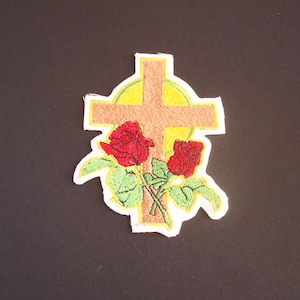 Könnte beinhalten: Gestickter Aufnäher mit einem Holzkreuz mit einem gelben Heiligenschein und zwei roten Rosen mit grünen Blättern.
