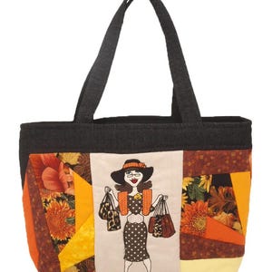 Op de afbeelding: Een patchwork-tas met een zwarte denim basis en kleurrijke stofpanelen met herfstthema's. De tas heeft een groot geborduurd beeld van een vrouw met een hoed en jurk die boodschappentassen vasthoudt.