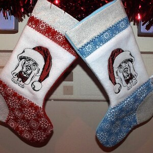 Puede incluir: Dos calcetines de Navidad con fondo blanco y patrones de copos de nieve rojos y azules. Cada calcetín presenta una imagen bordada de un perro sabueso de pelo corto con un gorro de Papá Noel.