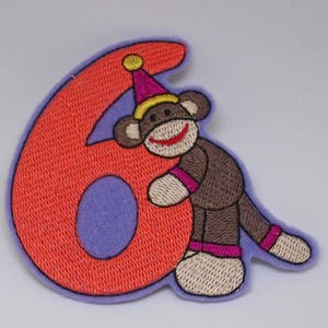 May include: An orange felt number six with a brown monkey wearing a pink and yellow party hat leaning against it.