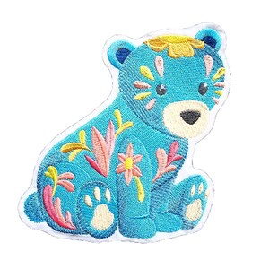 Könnte beinhalten: Ein blauer, gestickter Bügelaufkleber mit einem Bären mit floralen Details. Der Bär sitzt mit seinen Pfoten vor sich. Die Blumen sind gelb, rosa und orange.