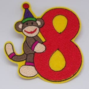 Puede incluir: Parche de hierro bordado con un mono marrón que lleva un gorro de fiesta morado y verde sosteniendo un número ocho rojo. El número ocho está rodeado de amarillo.