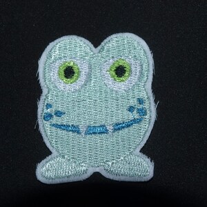Pode incluir: Um remendo de bordado de sapo azul claro com olhos verdes e boca azul. O sapo está sorrindo.