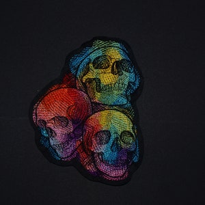 May include: Three embroidered skulls in a rainbow color scheme. The skulls are layered on top of each other, with the top skull being the largest. The skulls are all facing forward, with their eyes closed.