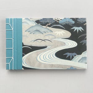 Puede incluir: Un diario rectangular con lomo turquesa y diseño de paisaje. La portada presenta un río que serpentea a través de una escena de montaña con árboles y pájaros, en tonos azules, grises y negros.