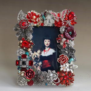 May include: A silver picture frame decorated with red, white, and black jeweled brooches, rhinestones, and flowers. The frame is filled with a portrait of a young girl with a dog.