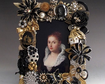 May include: A gold picture frame decorated with black and white jewels, rhinestones, and beads. The frame features a cameo of two people dancing and a black and white portrait of a woman.