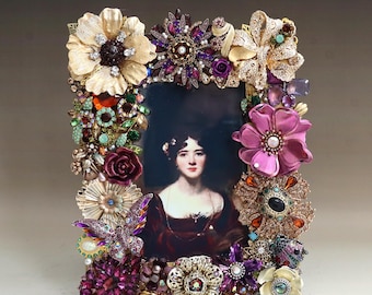 May include: A rectangular picture frame with a portrait of a woman. The frame is adorned with an array of decorative elements, including gold-toned flowers, purple and clear jewels, and various other embellishments. The portrait features a woman in a dark red dress.