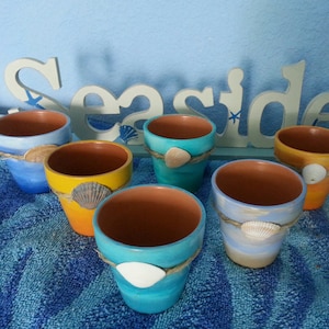 May include: Six small terracotta flower pots painted in shades of blue, yellow, and white with seashells glued to the outside. The pots are arranged on a blue and white striped towel. The word "Seaside" is written on a wooden sign behind the pots.